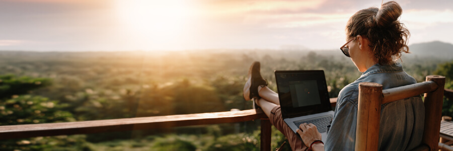 Woman sits with feet up on a balcony looking at an iPad,with a beautiful sunset view of the countryside