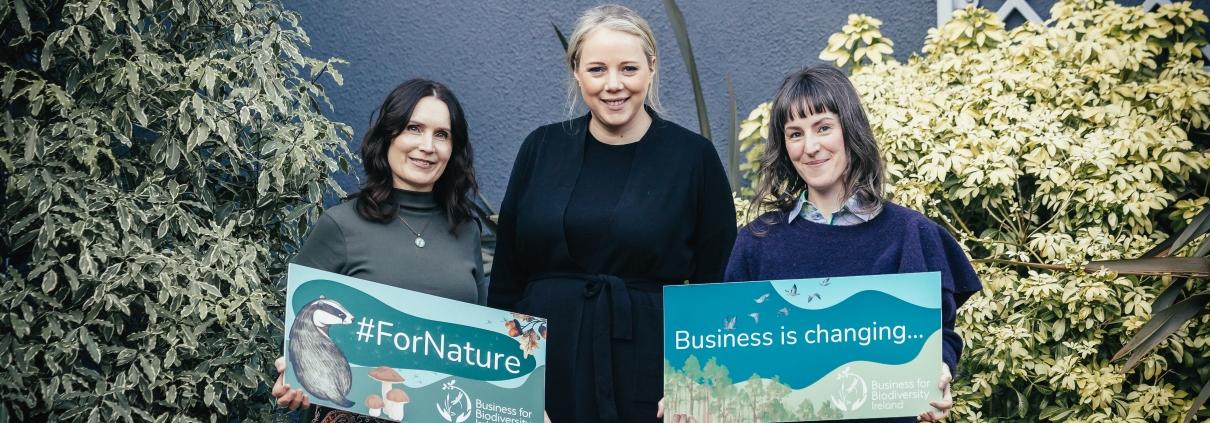 three women stand in a leafy garden, 2 holding signs with nature illustrations and slogans 'Business is changing...'and #ForNature