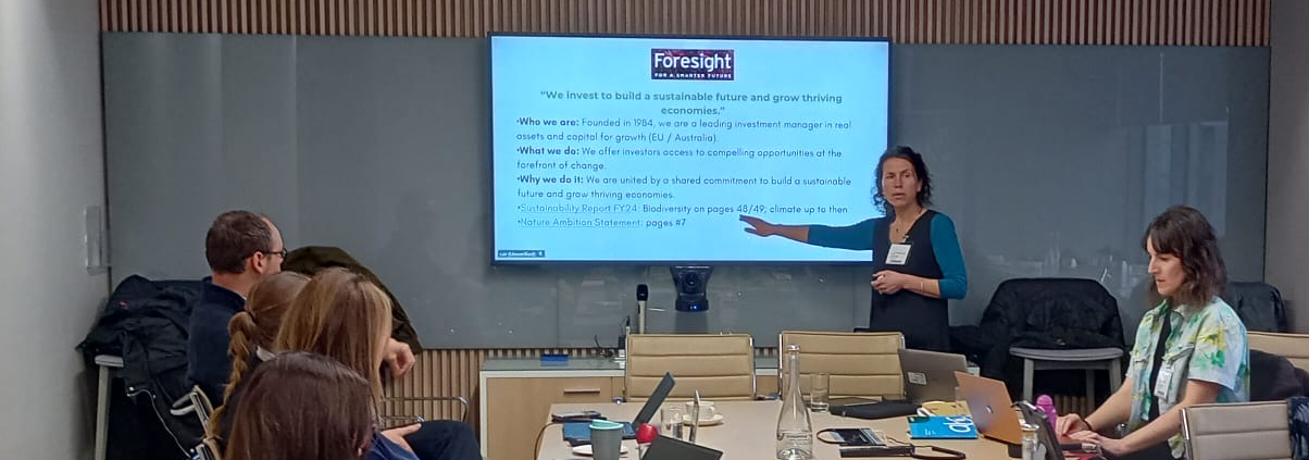 woman points at slide on screen at head of room while speaking to people at a meeting room table