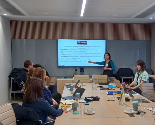 woman points at slide on screen at head of room while speaking to people at a meeting room table
