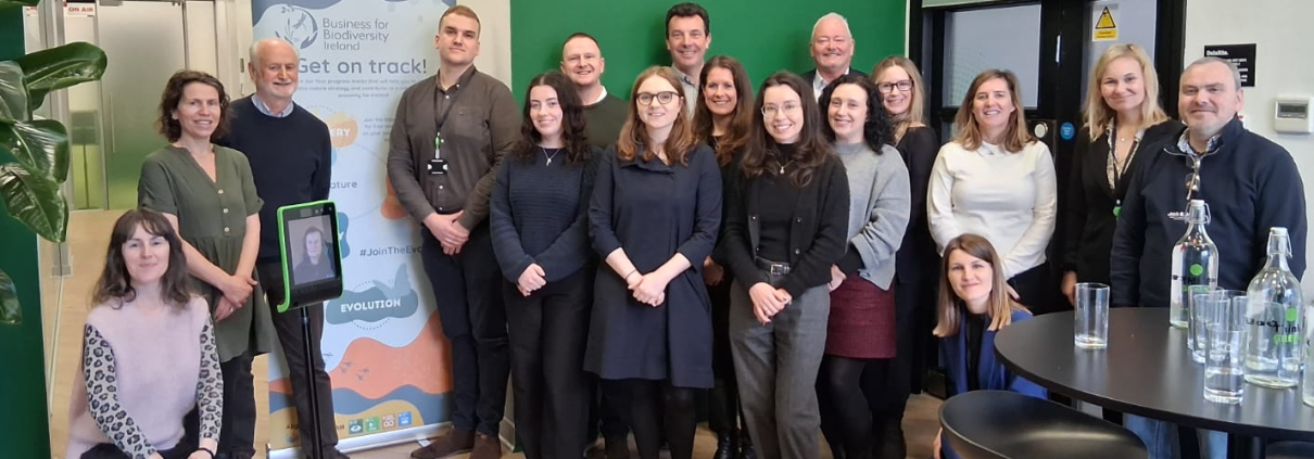 Group of people smiling in a function room with a green wall and pull-up poster, one person is dailling in on an iPad on a stand