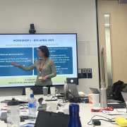 Woman stands at head of boardroom table and points at slide with blue boxes on screen