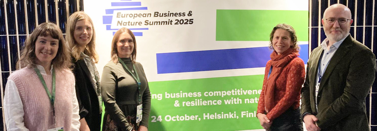 Four women and one man stand beside a poster in a confernce room, with logo for the European Business & Nature Summit in shades of green and blue