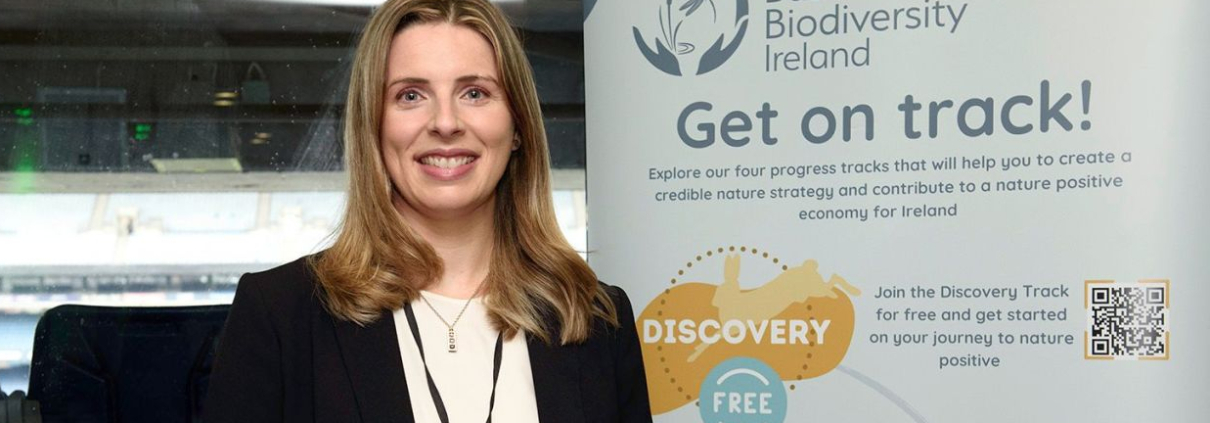 Smiling blonde woman wearing a lanyard and a black suit with a white top stands in front of a pull-up poster with logos, a stadium can be seen behind her through a window
