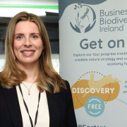 Smiling blonde woman wearing a lanyard and a black suit with a white top stands in front of a pull-up poster with logos, a stadium can be seen behind her through a window