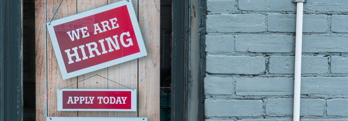 Picture of a door with a red sign with white text reading 'We are hiring'