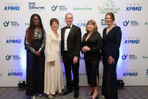 Four women and one man in evening wear at an awards ceremony, logos behind them
